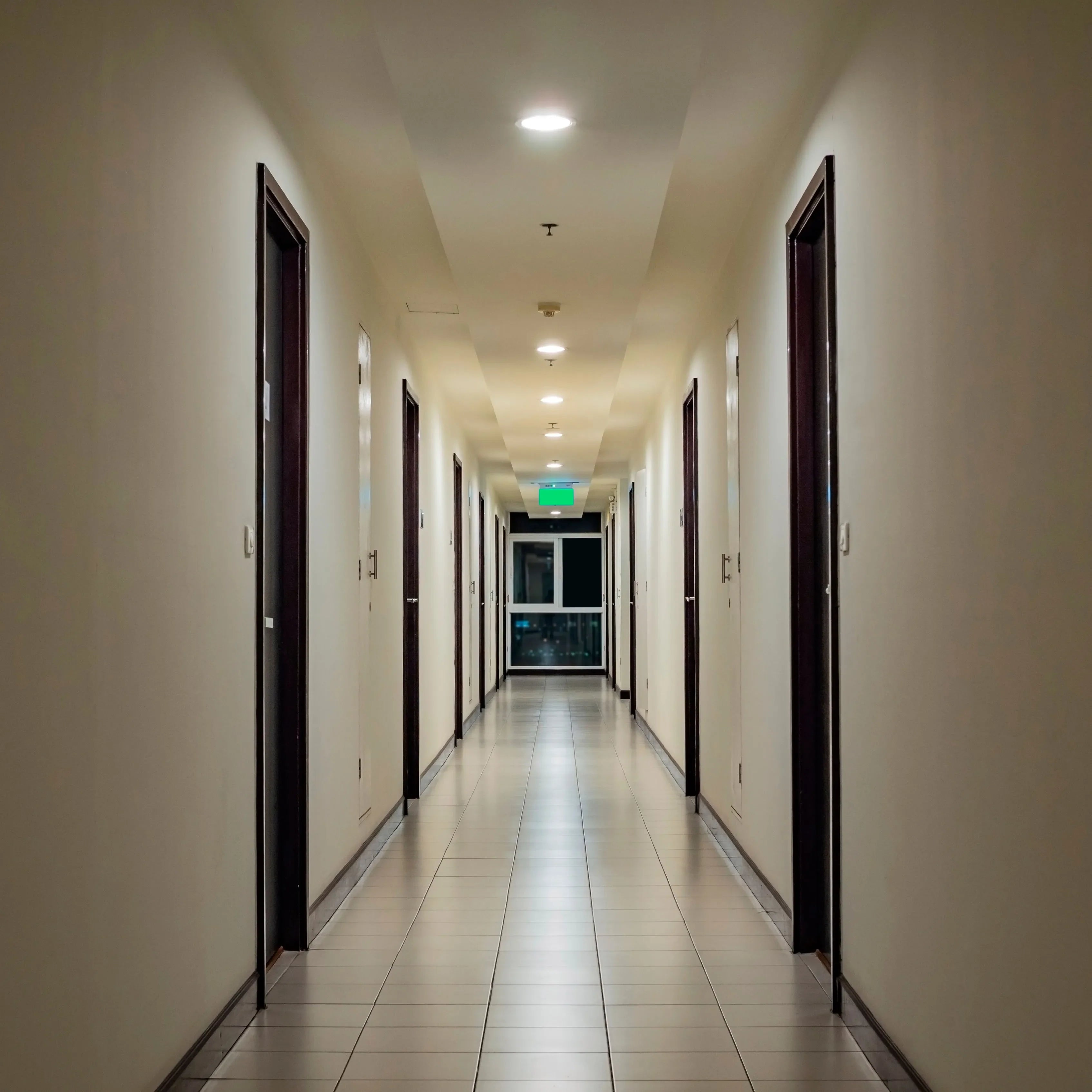 Recessed ceiling lights illuminate a long corridor with doors lining each side, and an exit sign at the end.