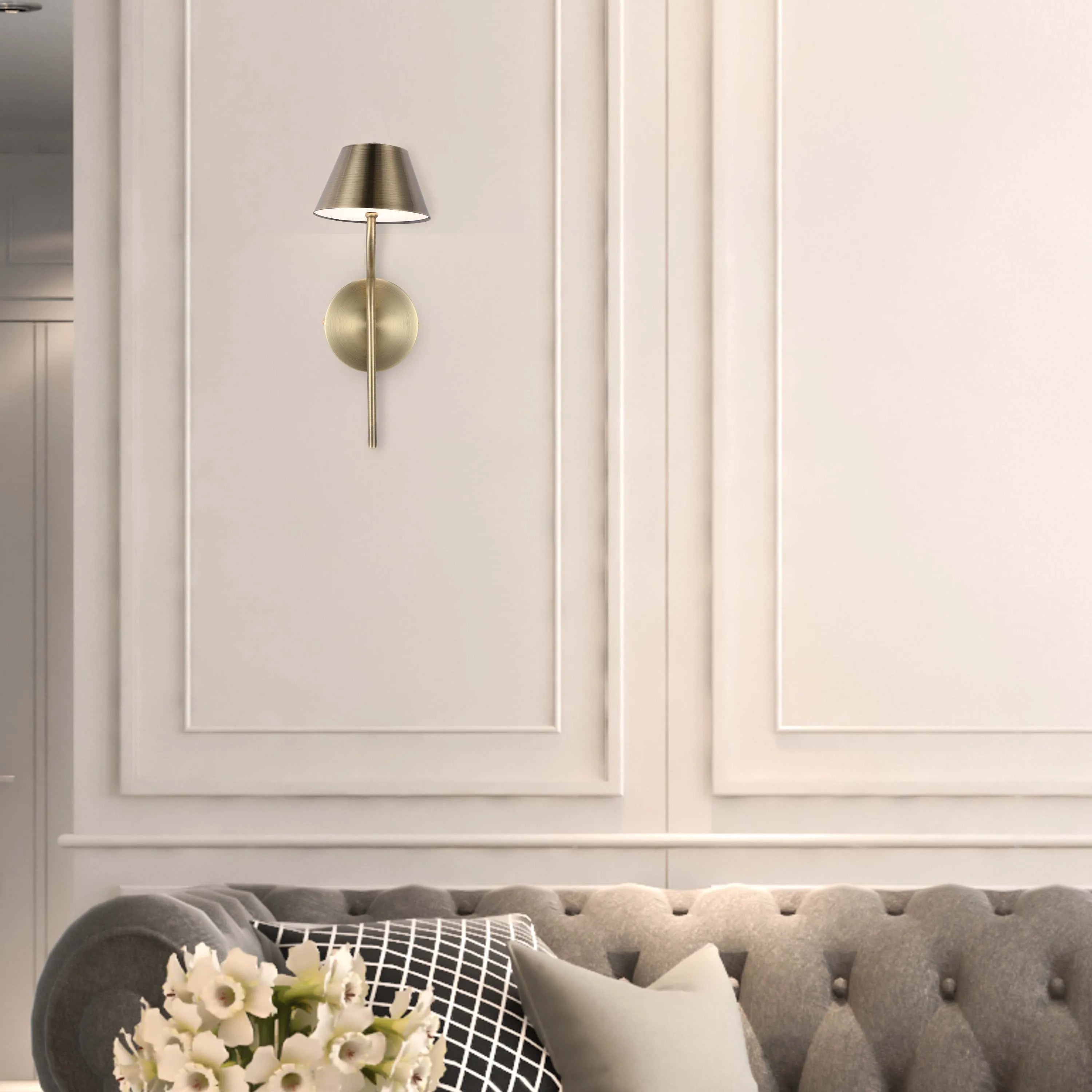 A modern gold wall sconce with a lampshade casts a warm glow against a panelled wall, illuminating a stylish sofa below.