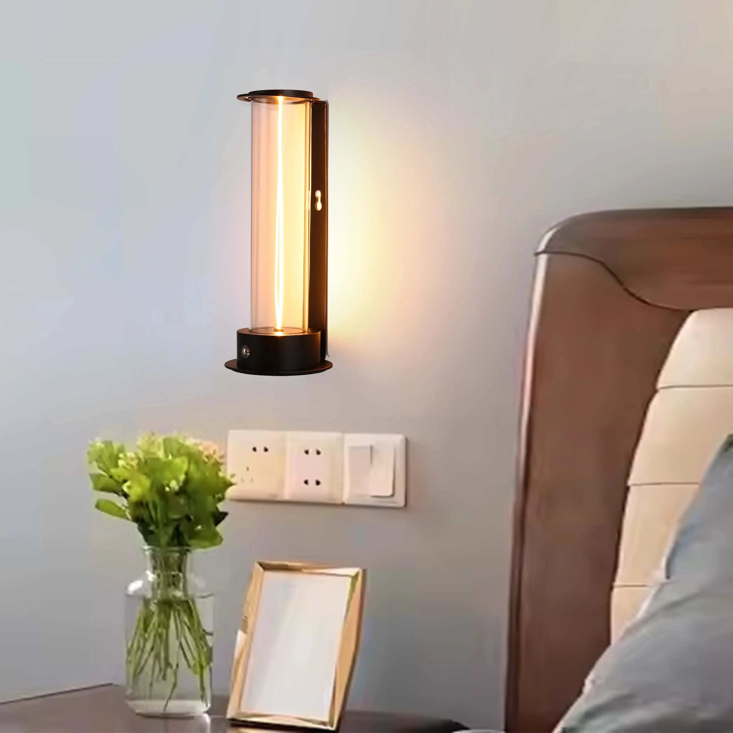 Modern wall light with cylindrical glass shade and warm glow, next to a bedside table with flowers and a photo frame.