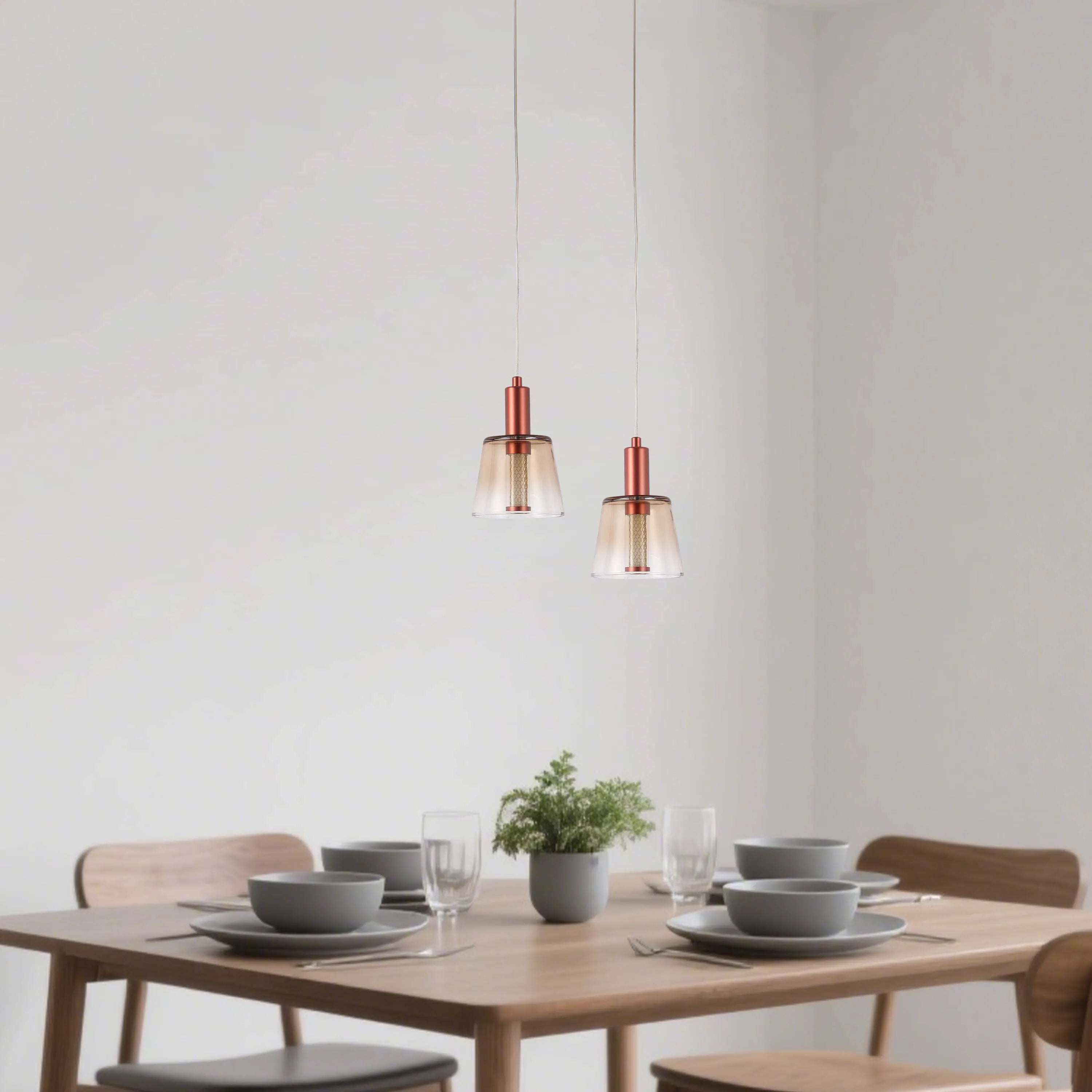 Two warm-toned pendant lights illuminate a wooden dining table set with minimalist tableware and a small potted plant.