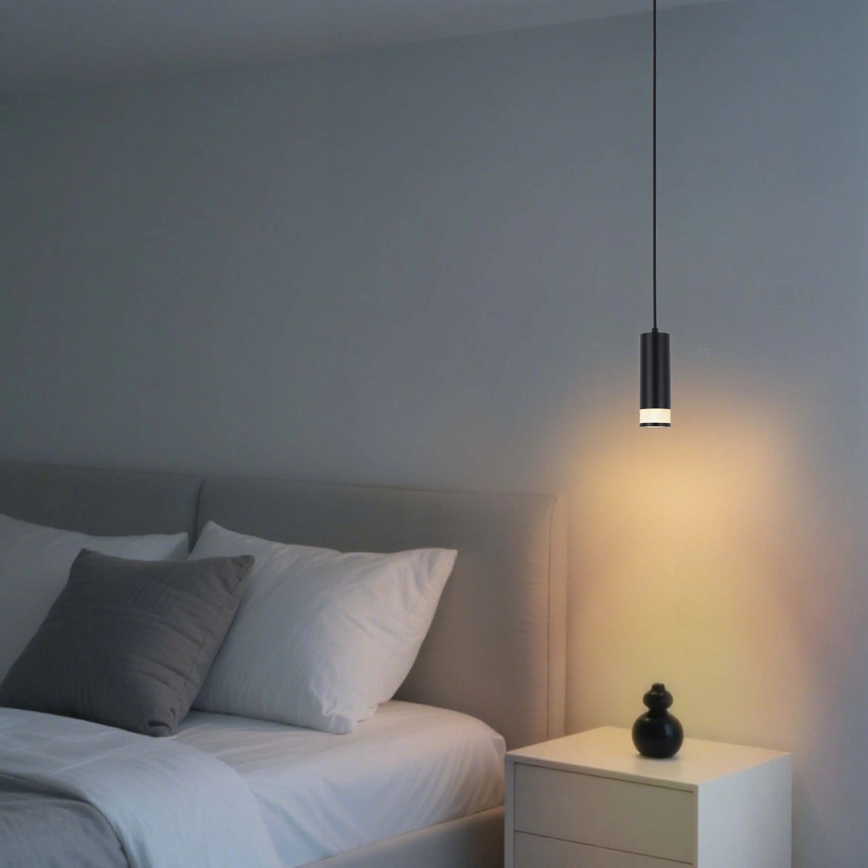 A modern cylindrical pendant light provides a warm glow beside a bed and bedside table, creating a cosy ambience.