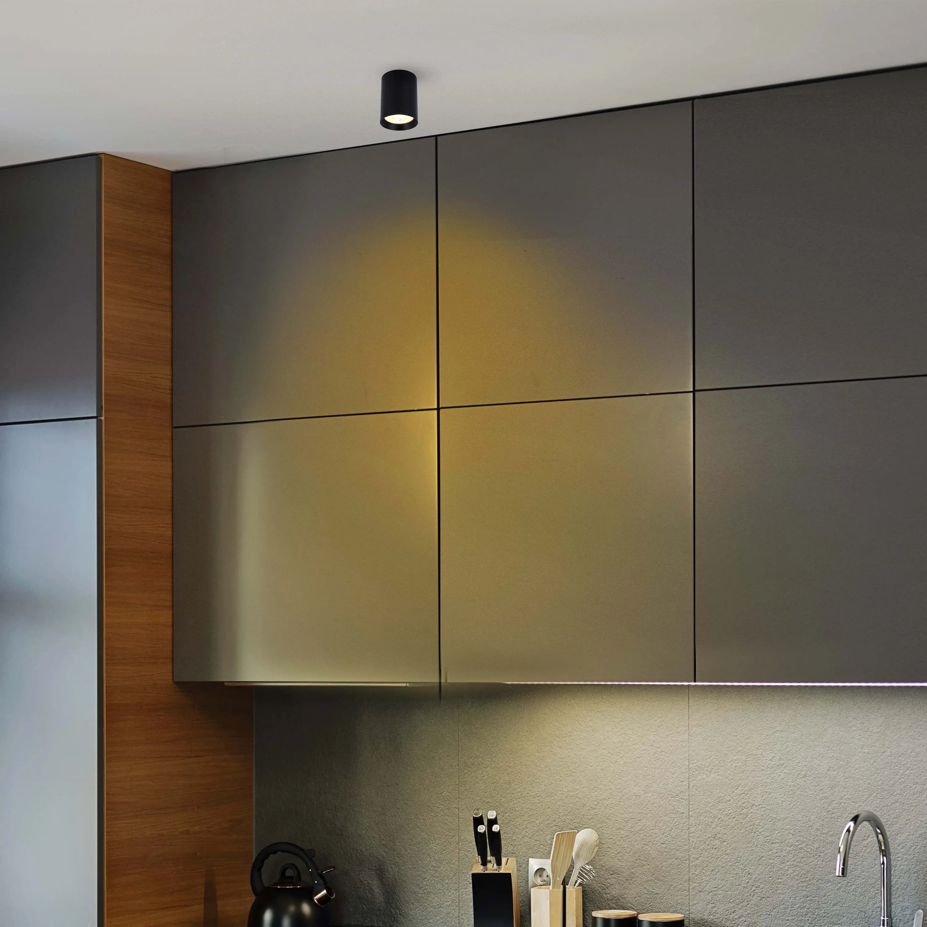 A modern kitchen with a spotlight on the ceiling and LED strip lighting under the cabinets. Dark grey cabinets provide overhead storage.