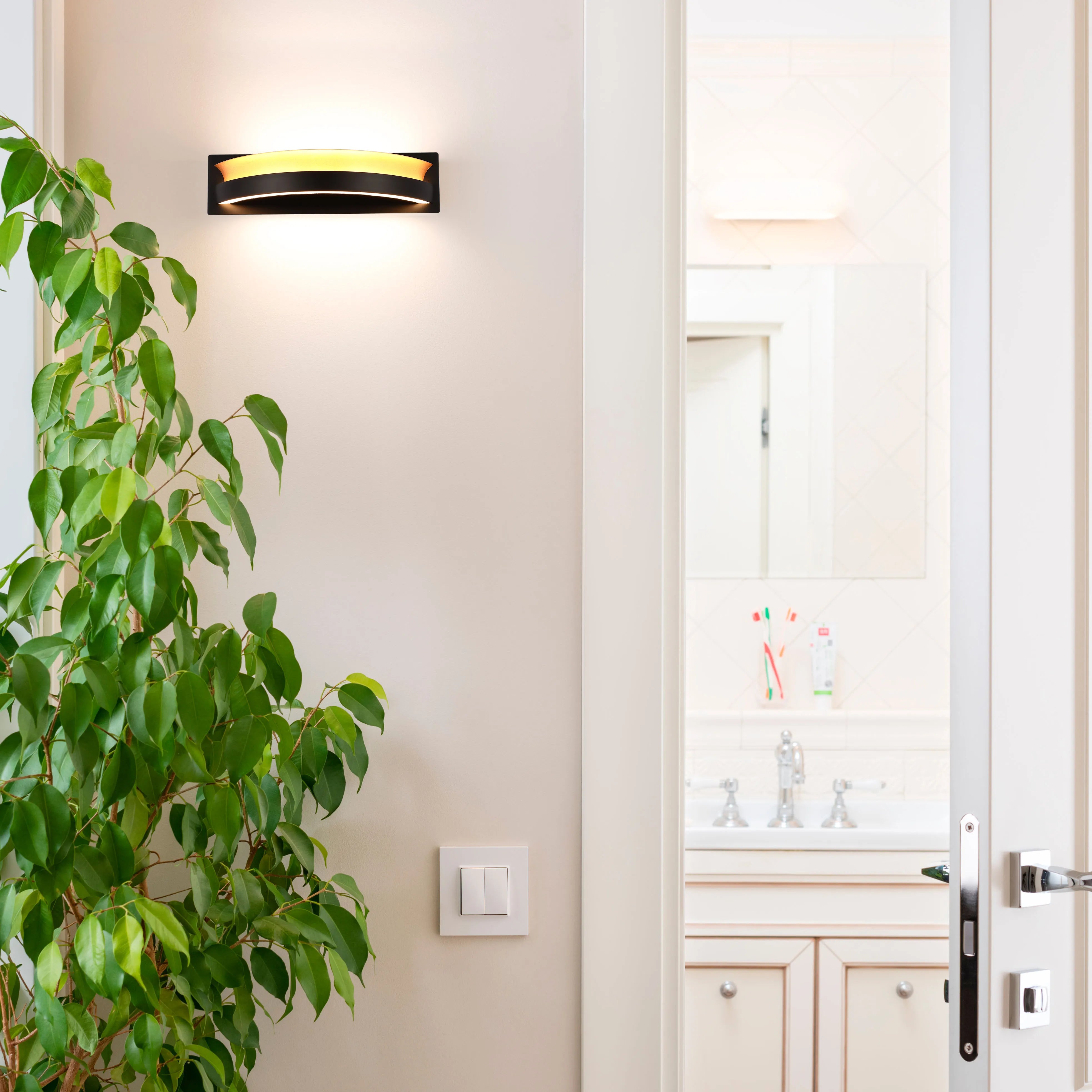 A contemporary wall light with a black surround emits a warm glow. Positioned above a light switch on a cream-coloured wall.