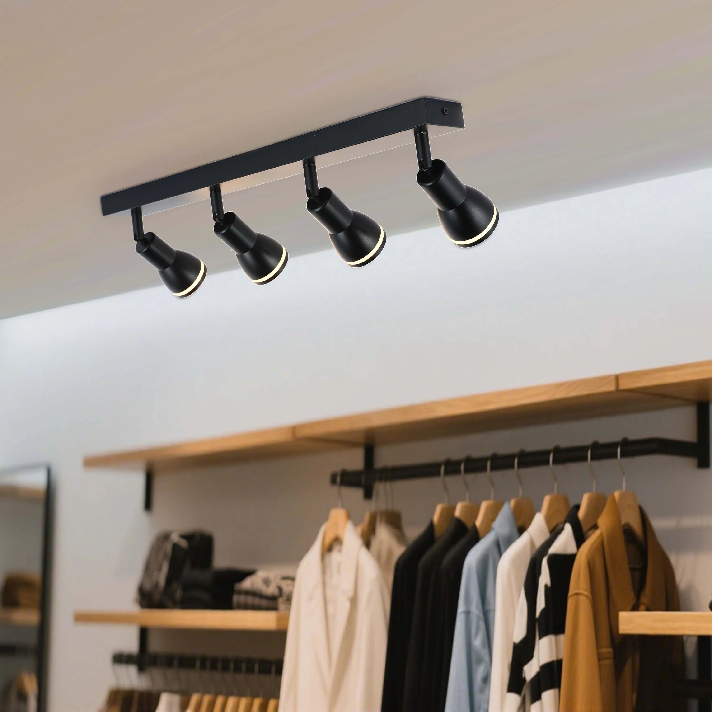 A black track lighting fixture with four adjustable spotlights, mounted on a white ceiling, illuminating clothes on hangers below.