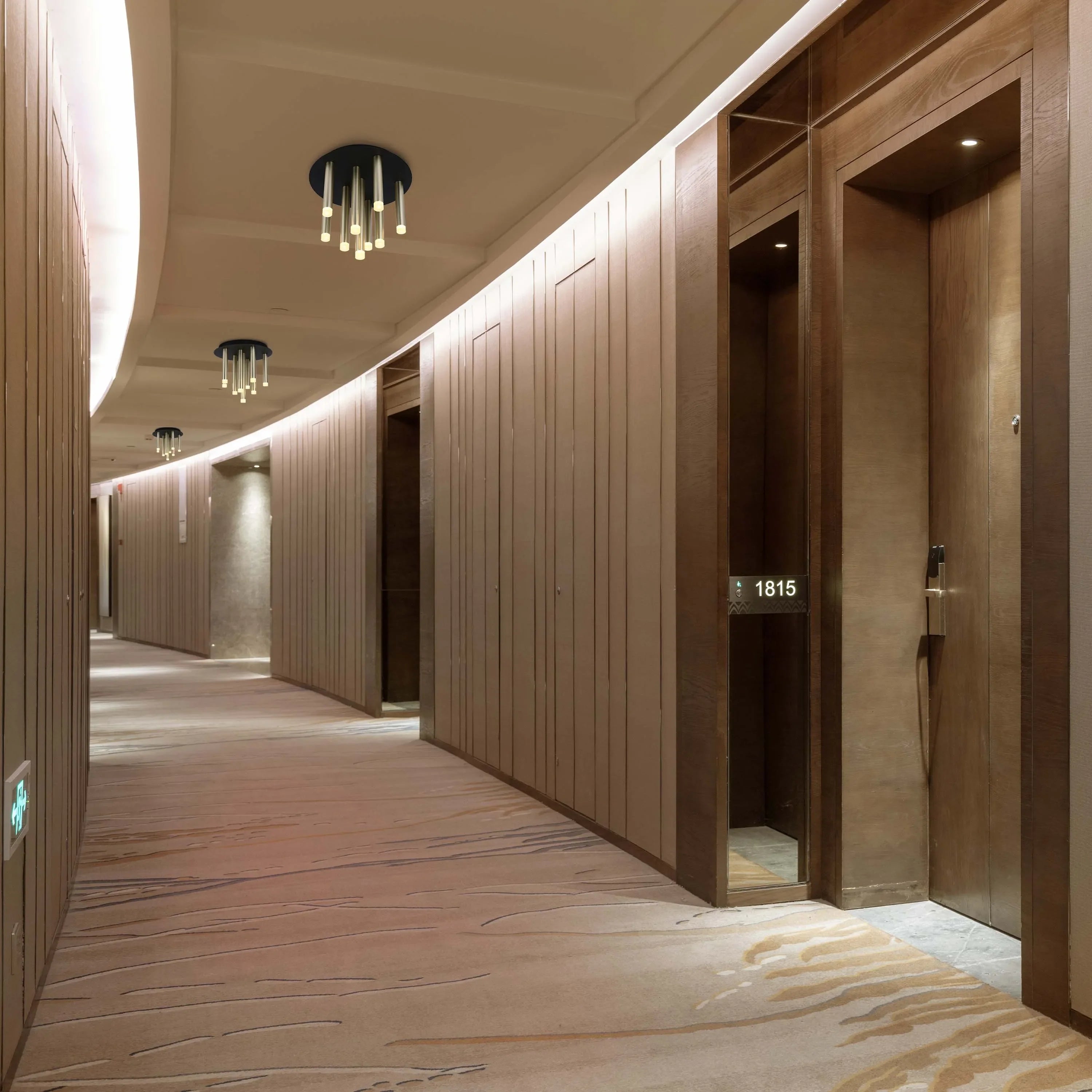 A hotel corridor is illuminated with ceiling lights and recessed spotlights above each room door, creating a warm and inviting ambience.