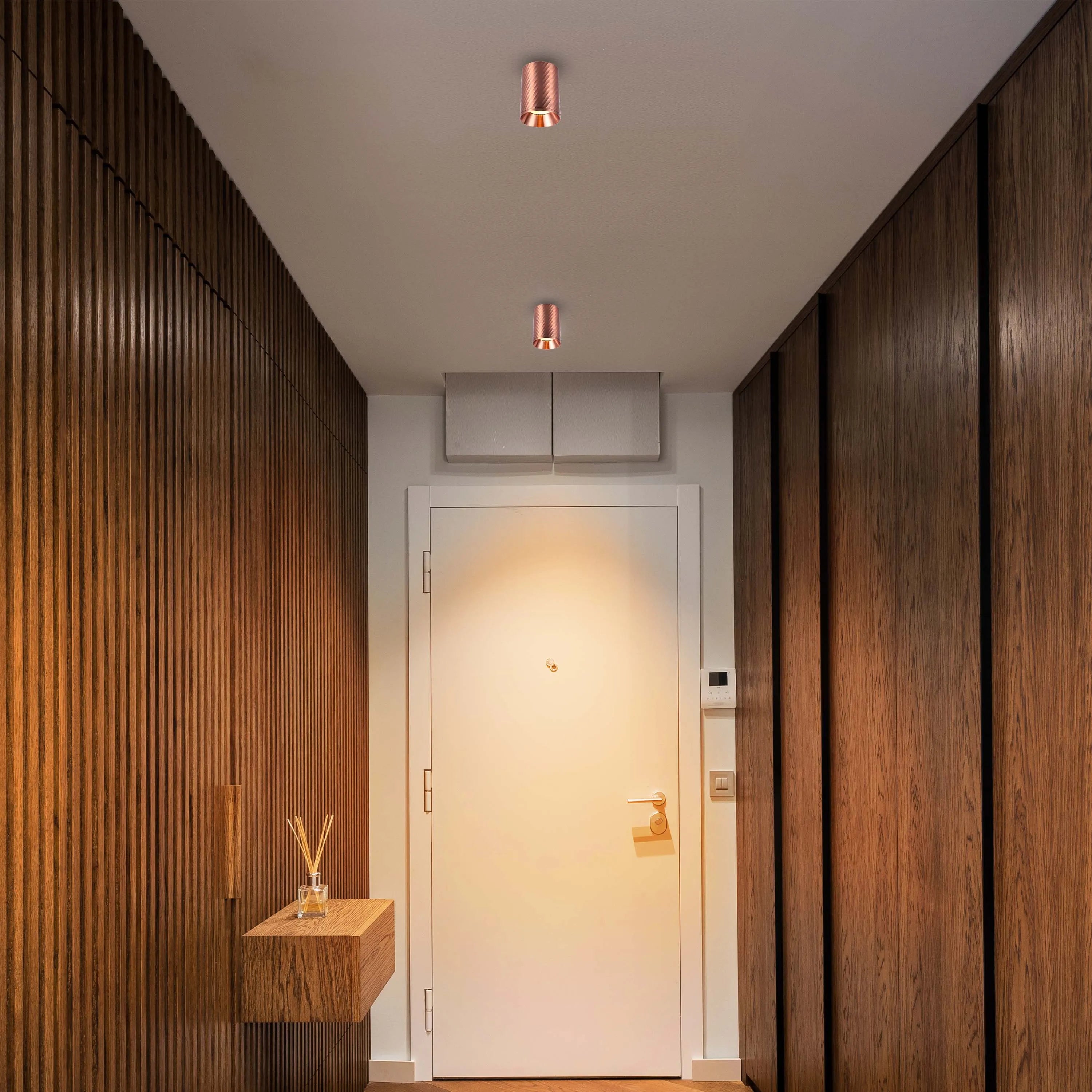 A modern hallway is illuminated by two copper, cylindrical ceiling lights, creating a warm and inviting ambience.