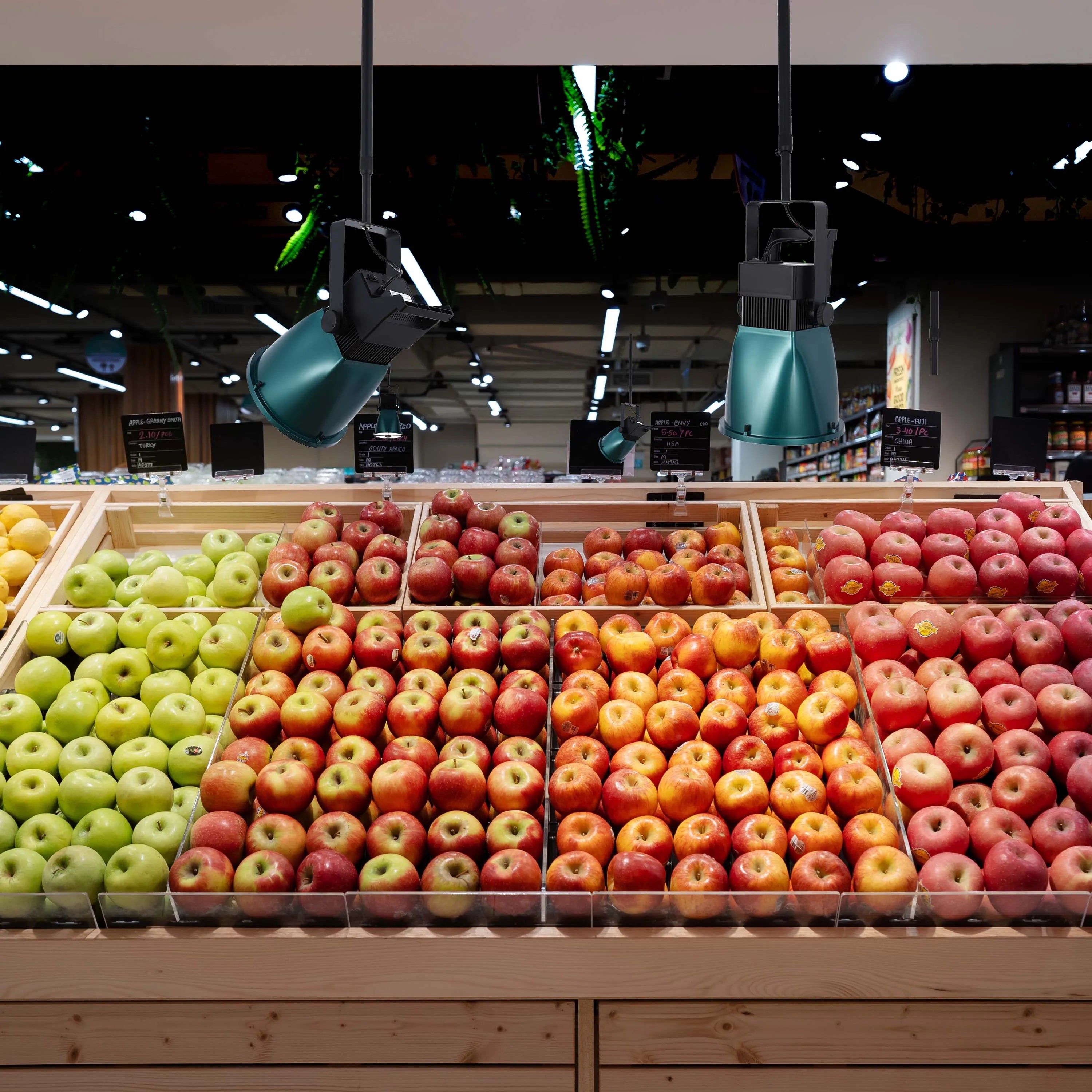 Track lighting illuminates displays of fresh apples in a supermarket, creating bright, focused illumination for product presentation.