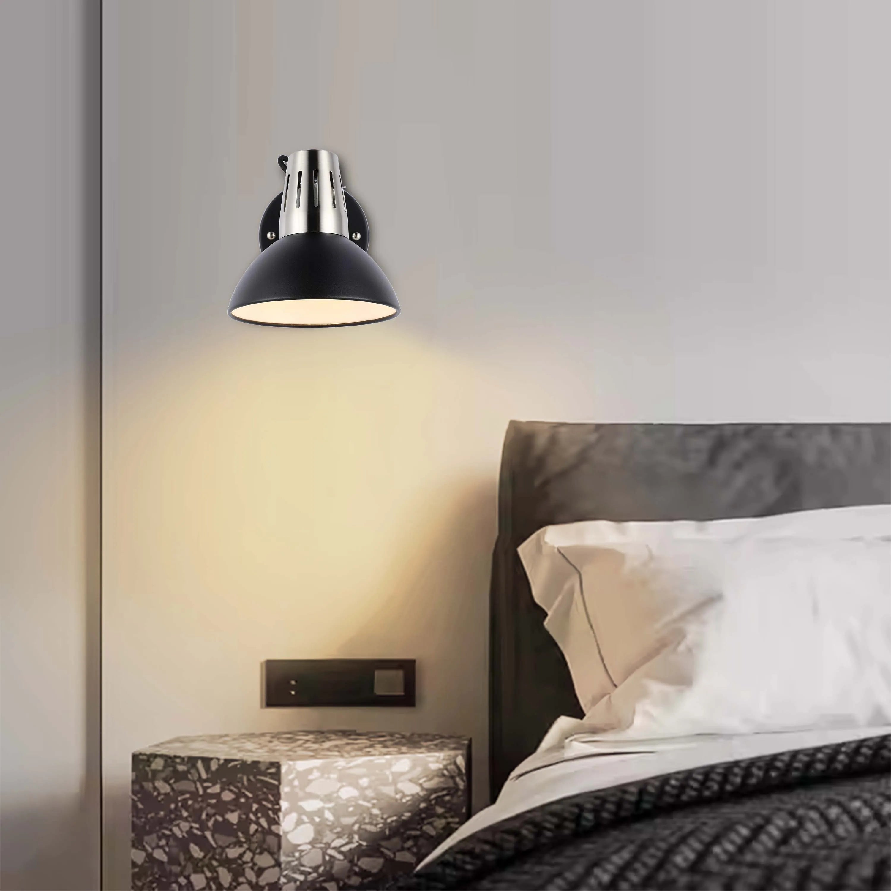 A modern wall light with a black dome shade and silver detailing, casting a warm glow in a contemporary bedroom setting.