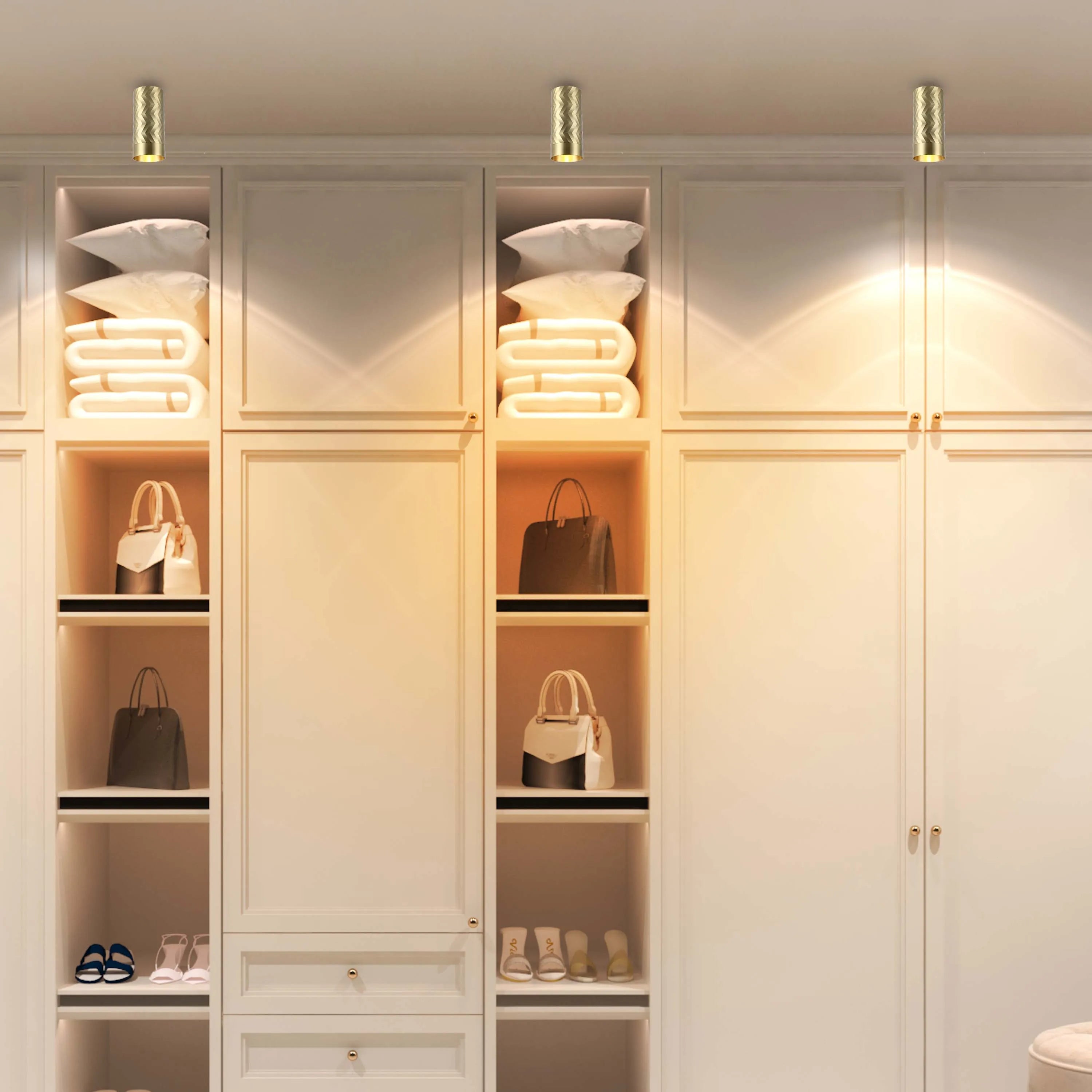 Modern, gold cylindrical ceiling lights illuminate a bright wardrobe. The warm glow emphasises the cream and beige tones of the closet.