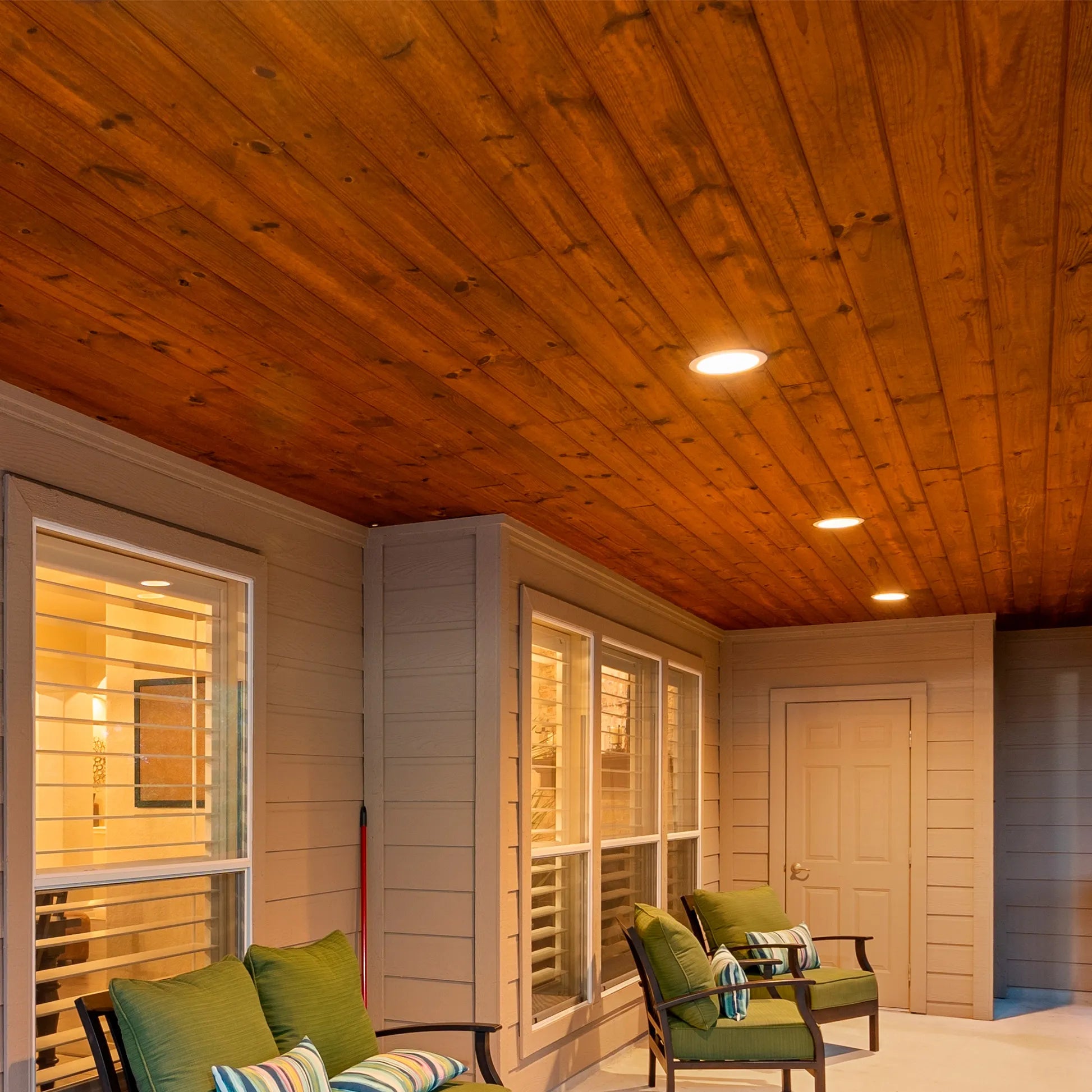 Warm lighting from recessed ceiling lights illuminates an outdoor porch with wooden ceiling and neutral coloured walls.