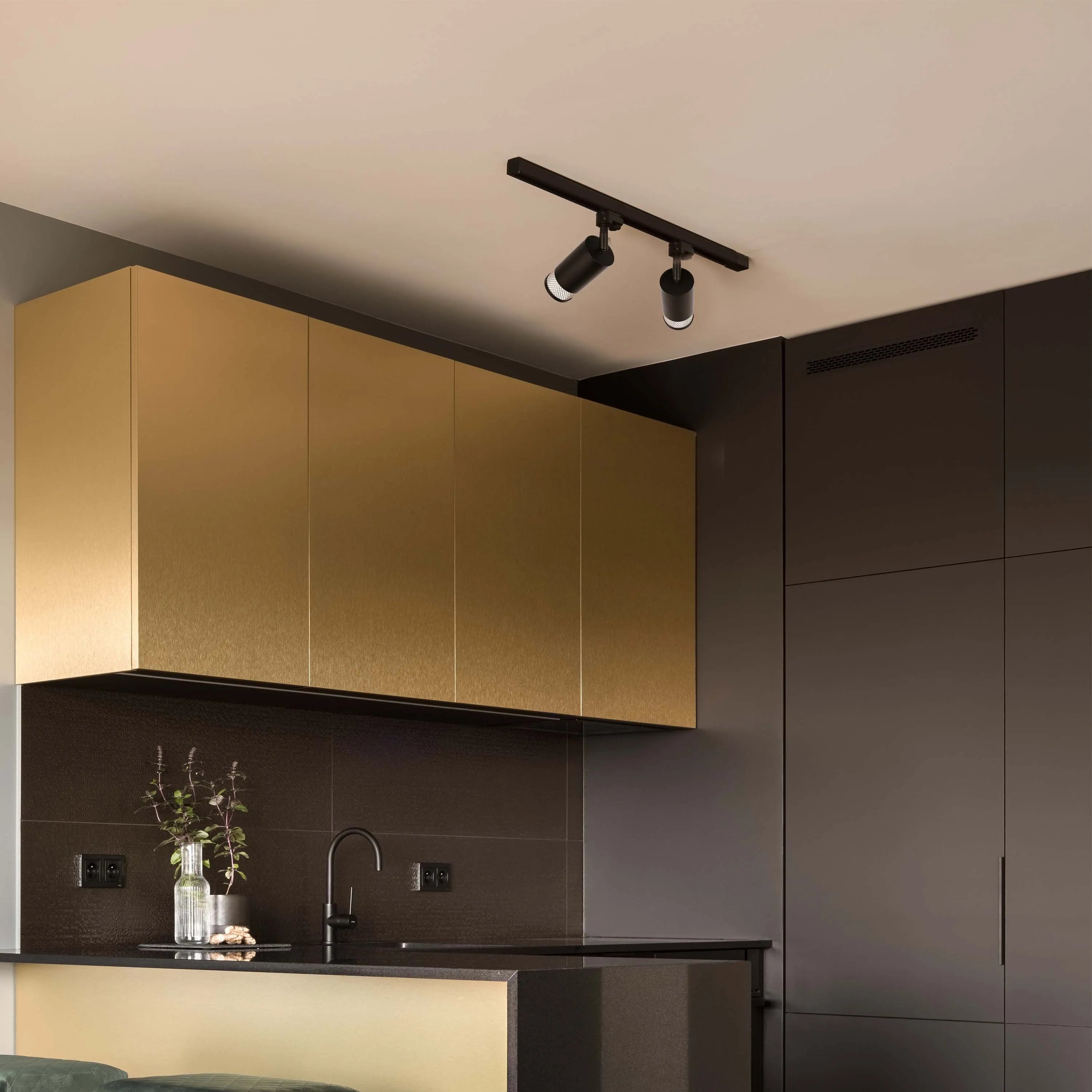 A black track light fixture with two spotlights illuminates the modern kitchen. The lights are focused on the gold cabinetry below.