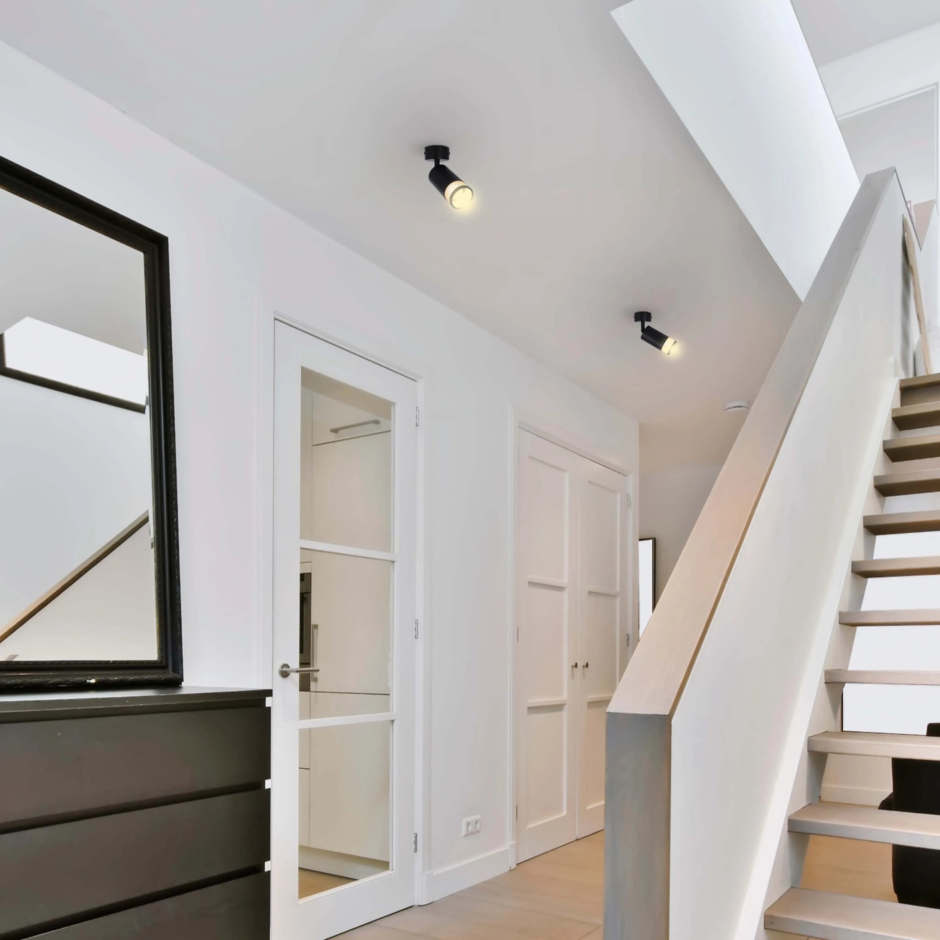 Recessed spotlights illuminate a hallway with white walls, a wood floor, a dark chest of drawers, and a staircase with a white banister.