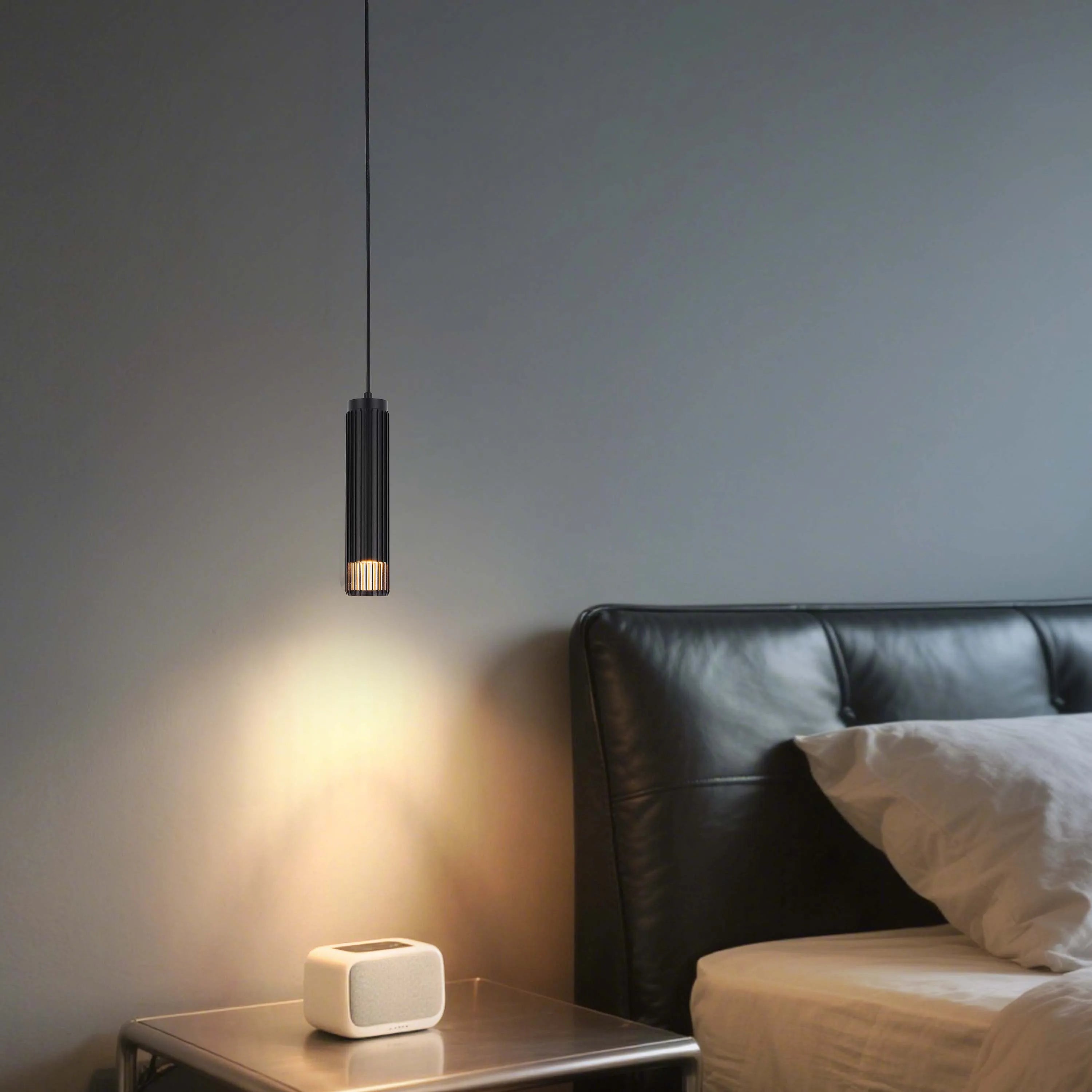 A modern black pendant light casts a warm glow in a bedroom setting, illuminating a bedside table and adjacent bed.