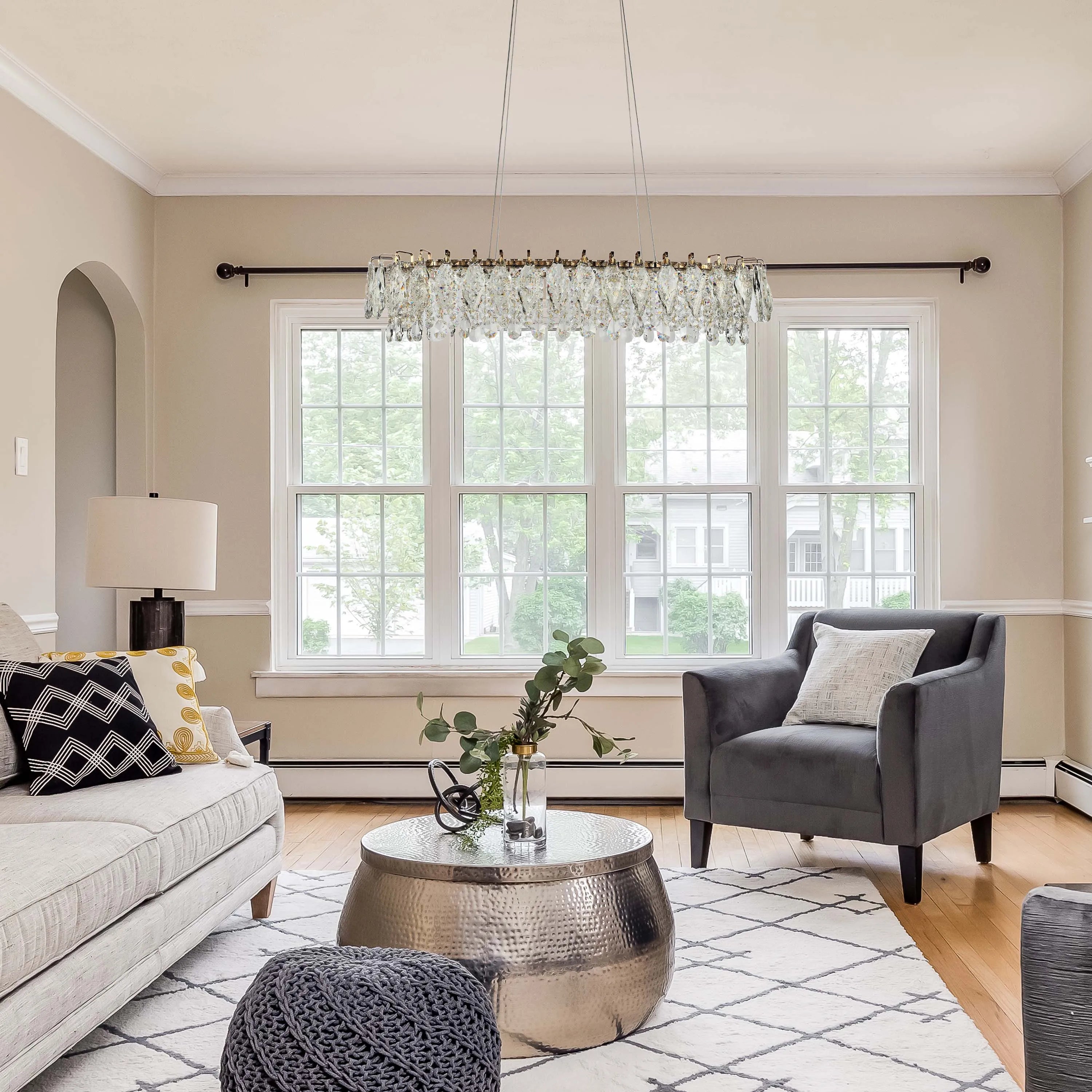 A crystal chandelier hangs in a living room, casting a soft, elegant glow. A table lamp provides additional lighting.