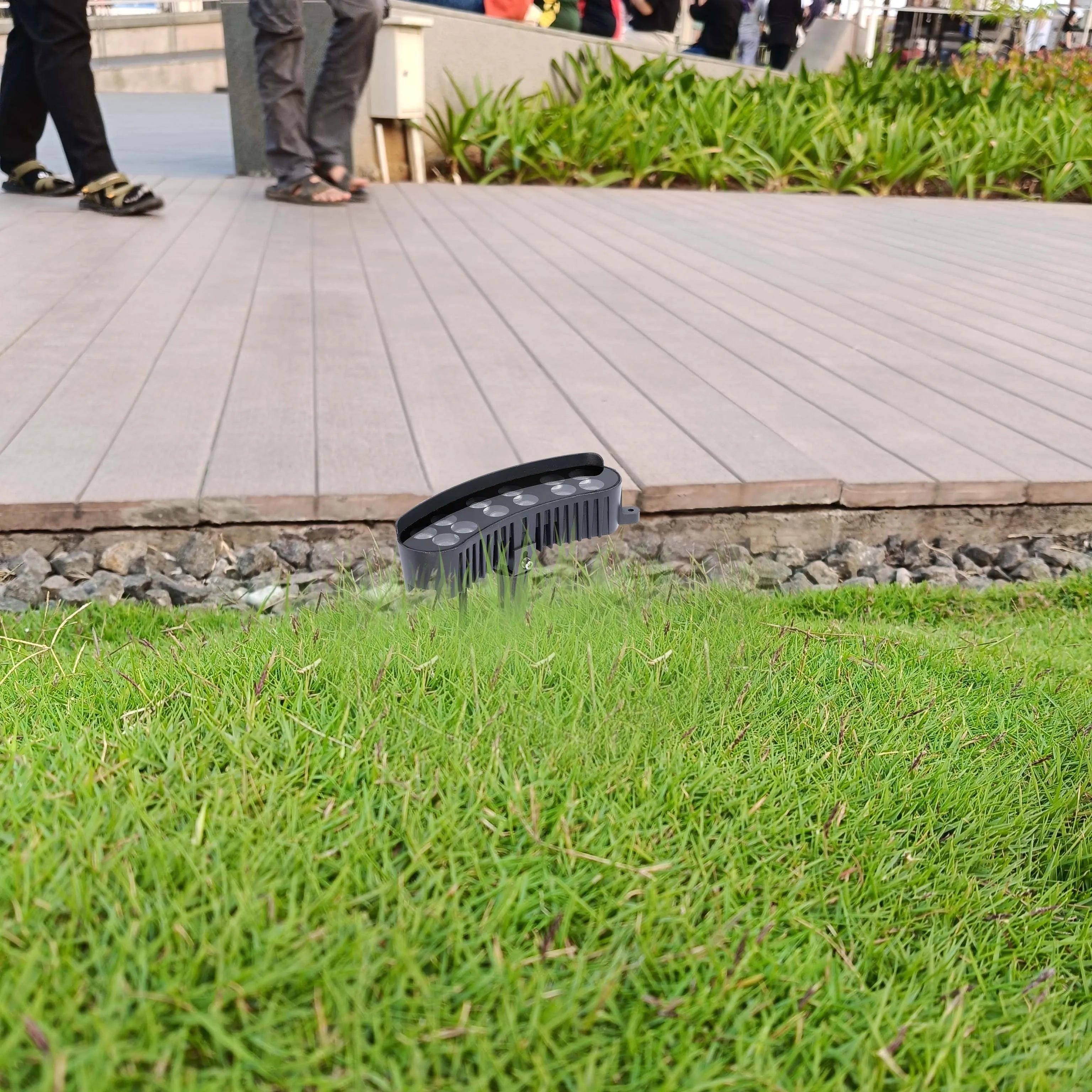 Outdoor LED ground light on a patch of green grass, with a wooden walkway and some people in the background.