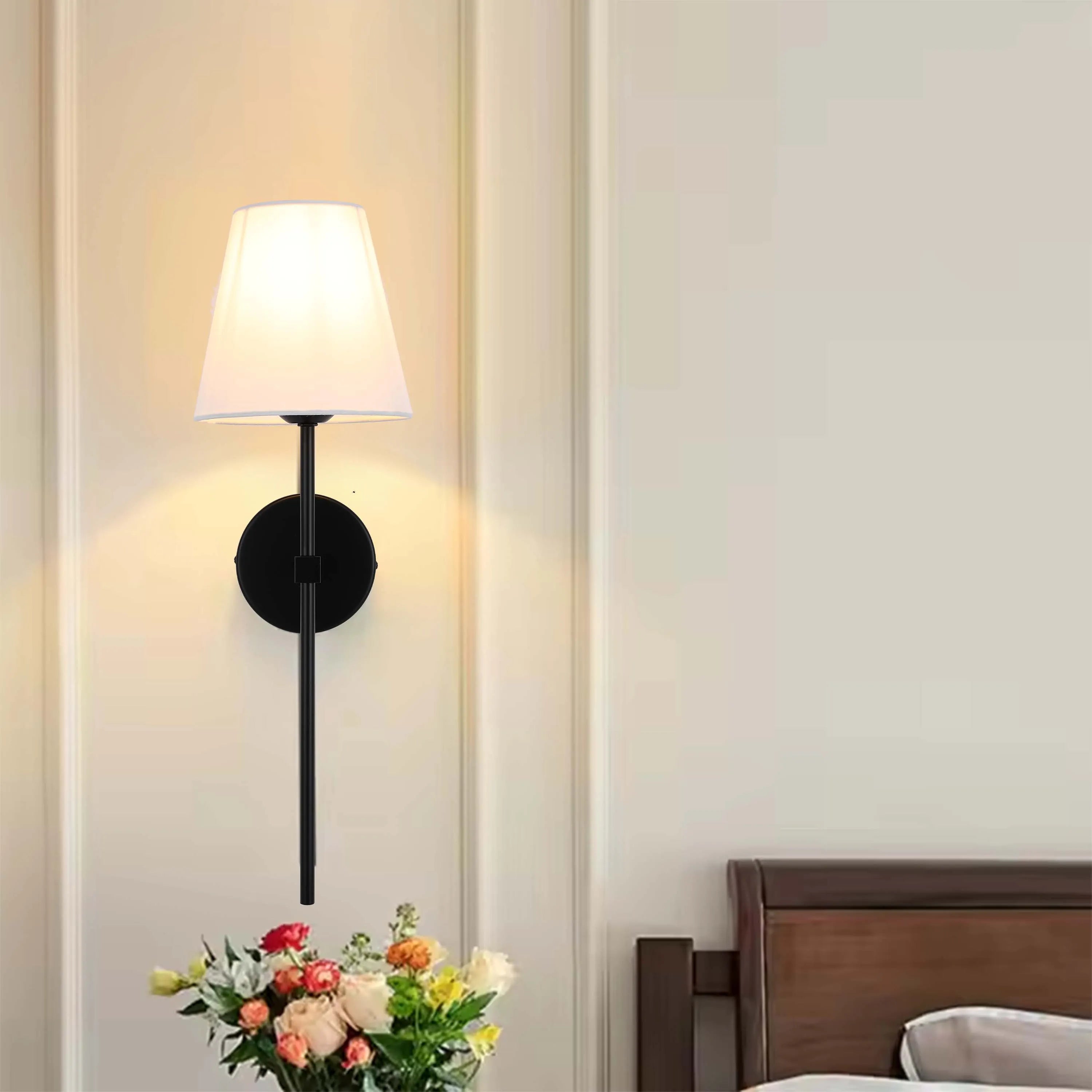 Wall-mounted lamp with a white lampshade & black fixture, casting a soft glow on the cream-coloured wall. A vase of flowers sits below.
