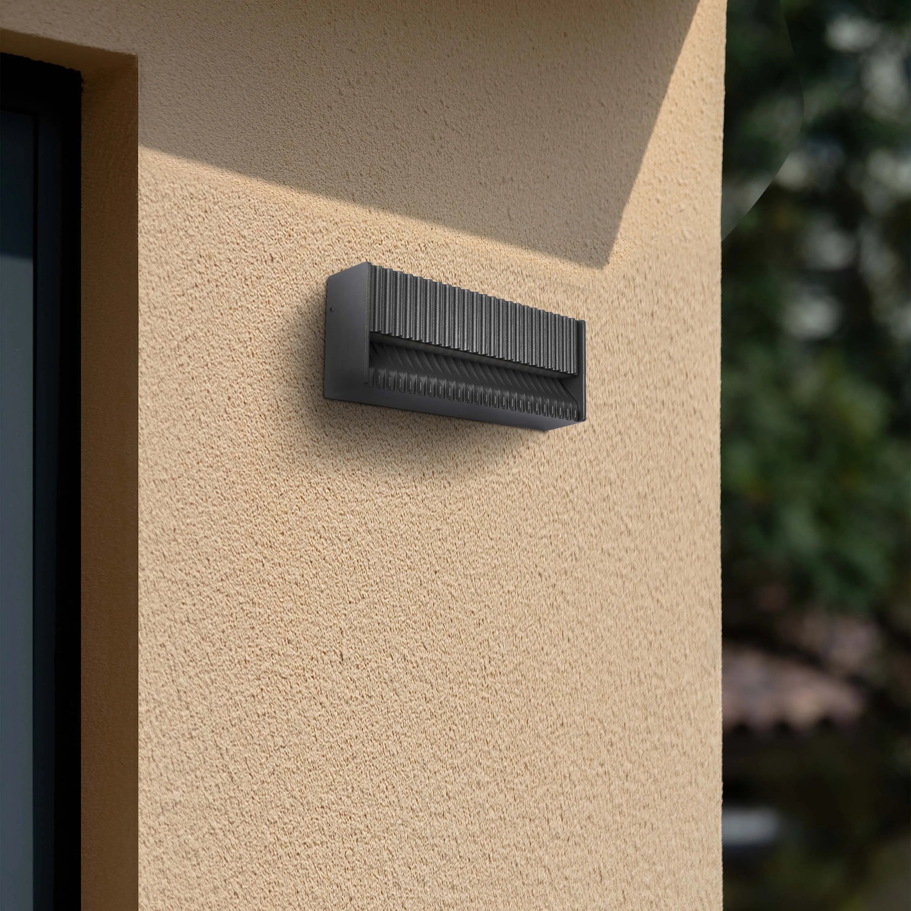 Modern outdoor wall light with a textured finish, casting shadows that emphasise its grooved design. Bright, natural sunlight floods the scene.