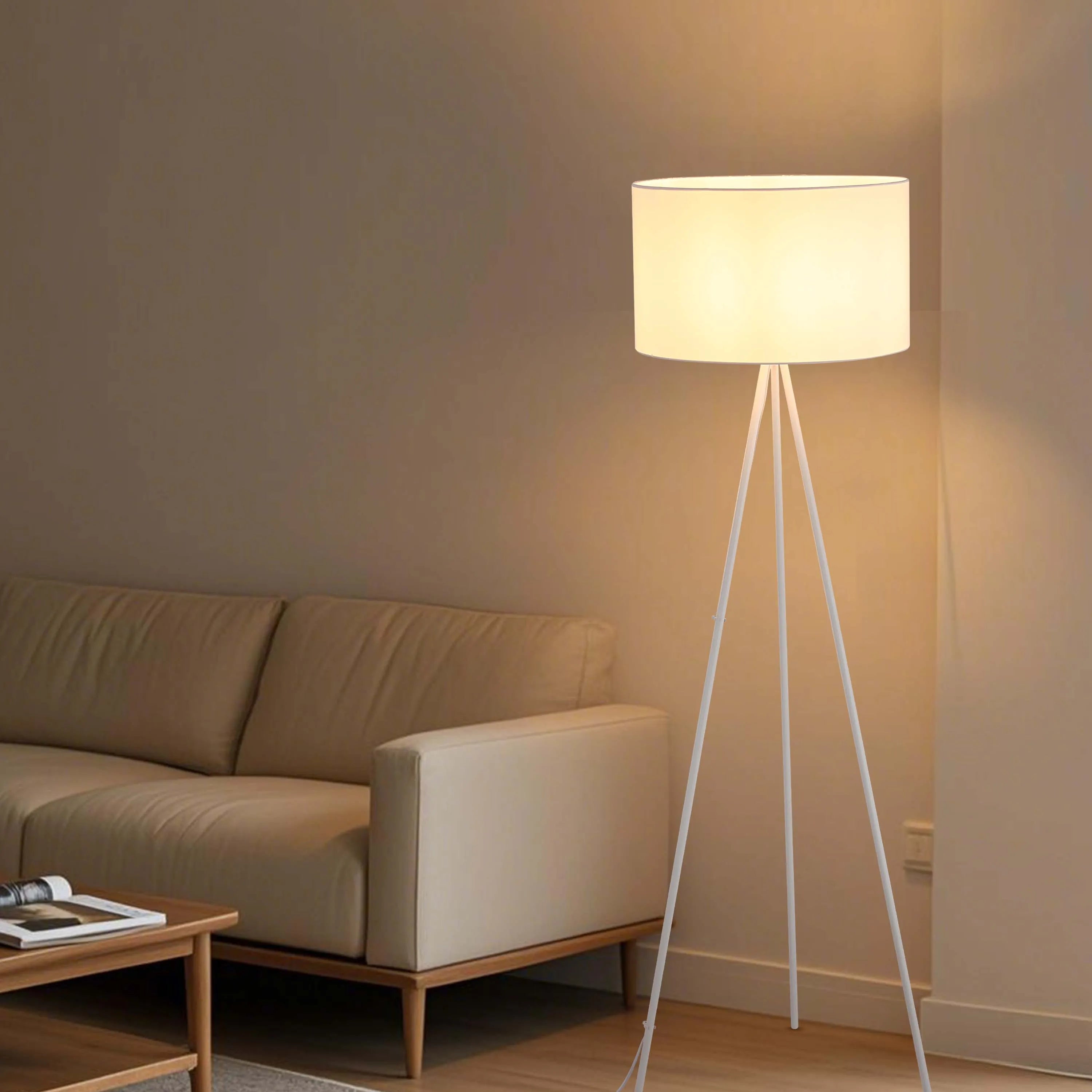 A modern floor lamp with a white shade atop a sleek, white tripod base, emitting a warm, inviting glow in a neutral living space.